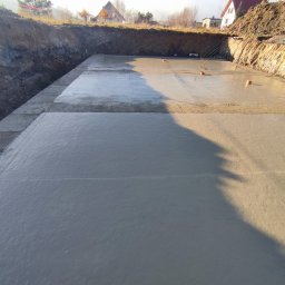 Perbud - Świeżo wylany fundament pod budynek jednorodzinny, widoczne szalunki i wystające pręty zbrojeniowe, teren przygotowany pod dalsze prace budowlane.