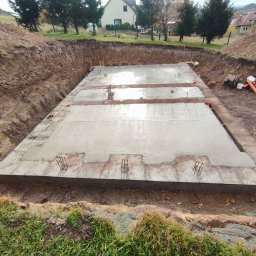 Perbud - Wylany fundament pod budynek, widoczne wystające pręty zbrojeniowe, teren wykopu z ziemią i fragment zabudowy w tle.