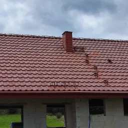 Czerwony dach z dachówki ceramicznej z kominem i zabezpieczeniami przeciwśniegowymi na surowym budynku z białych bloczków.