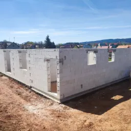 Wzniesione ściany parteru z betonu komórkowego na fundamencie z widocznymi otworami okiennymi i drzwiowymi na tle krajobrazu miejskiego.