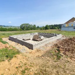 Wzniesione ściany fundamentowe z bloczków betonowych na działce, z widocznym szkieletem domu w tle, częściowo obłożonym izolacją.
