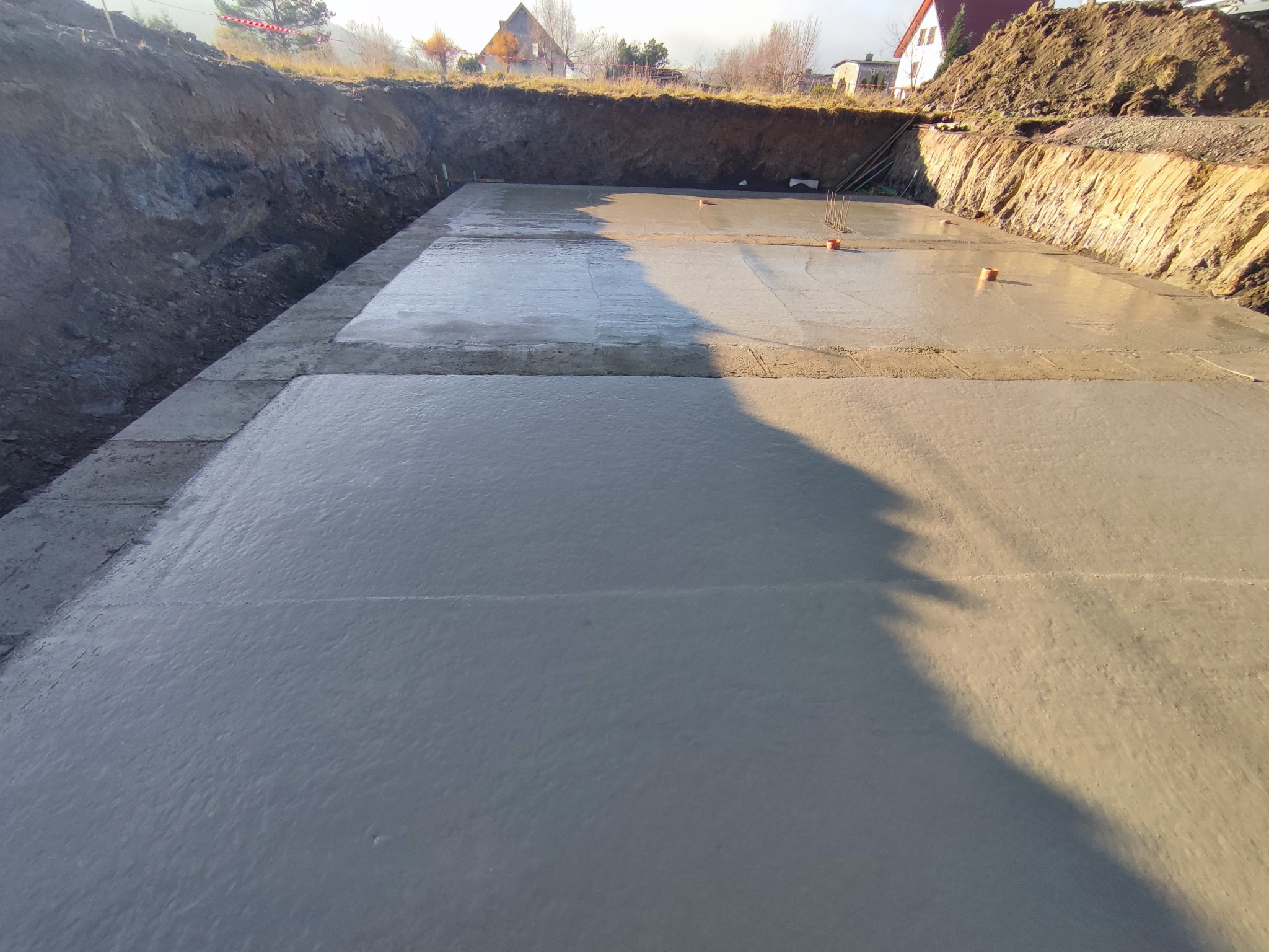 Świeżo wylany fundament pod budynek jednorodzinny, widoczne szalunki i wystające pręty zbrojeniowe, teren przygotowany pod dalsze prace budowlane.
