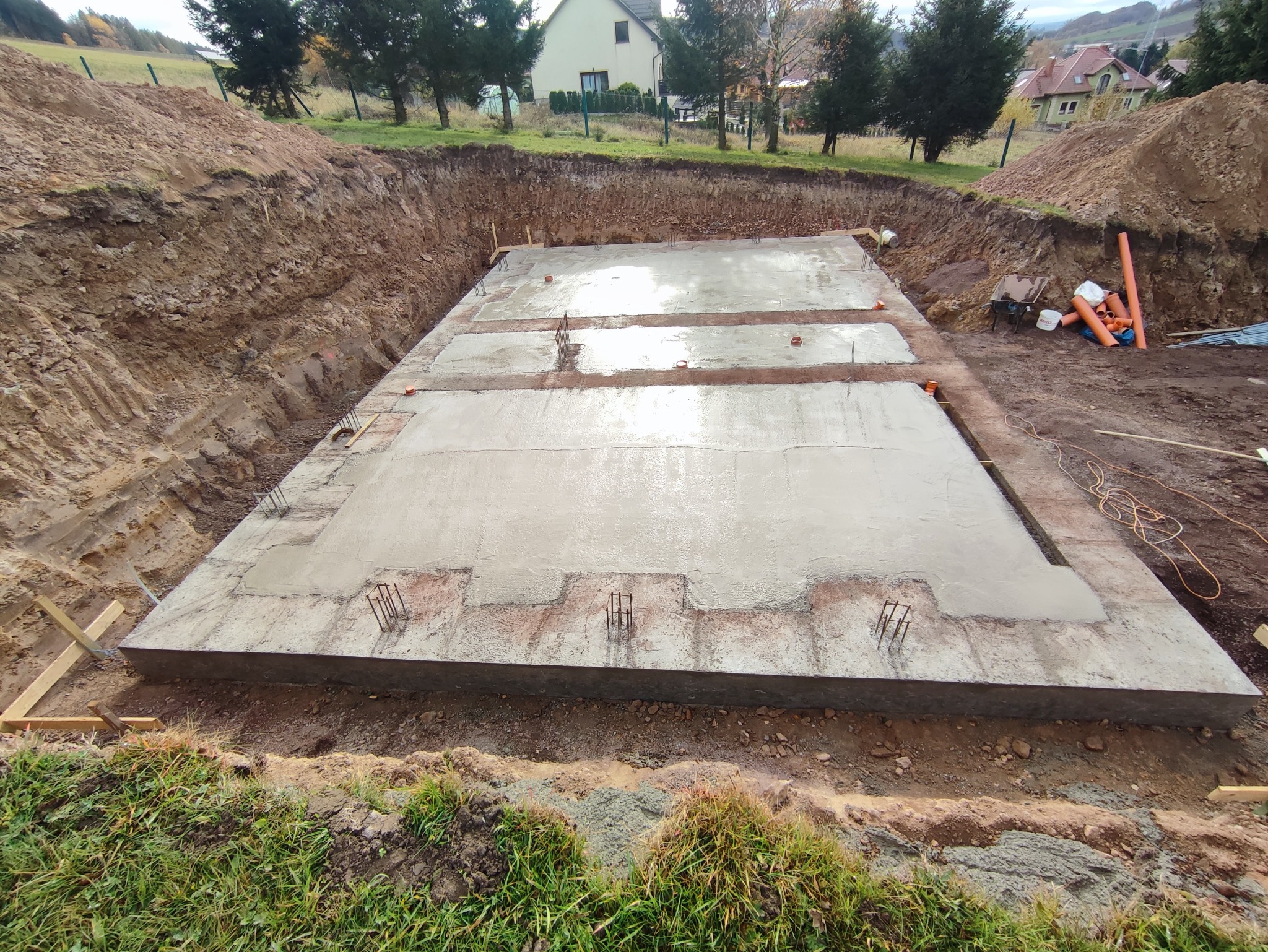 Wylany fundament pod budynek, widoczne wystające pręty zbrojeniowe, teren wykopu z ziemią i fragment zabudowy w tle.