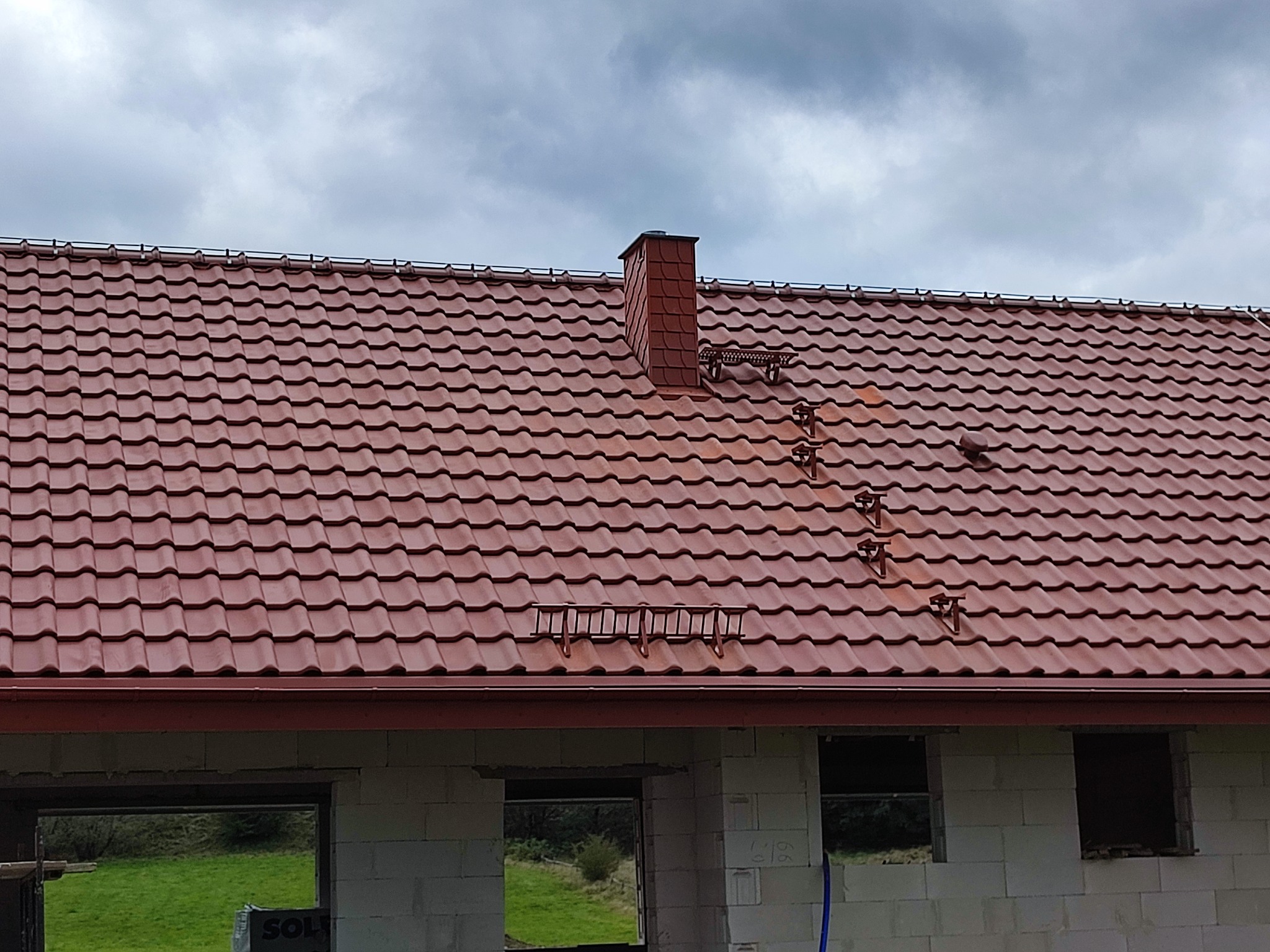 Czerwony dach z dachówki ceramicznej z kominem i zabezpieczeniami przeciwśniegowymi na surowym budynku z białych bloczków.