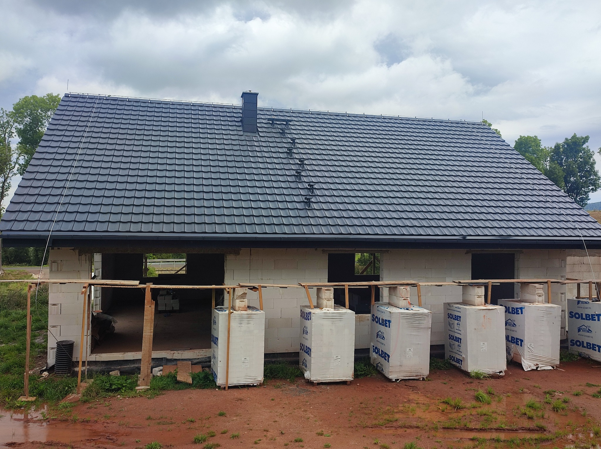 Niedokończony dom z białego betonu komórkowego, z szarym dachem z dachówki i kominem, palety z materiałami budowlanymi Solbet na błotnistym podłożu przed budynkiem.