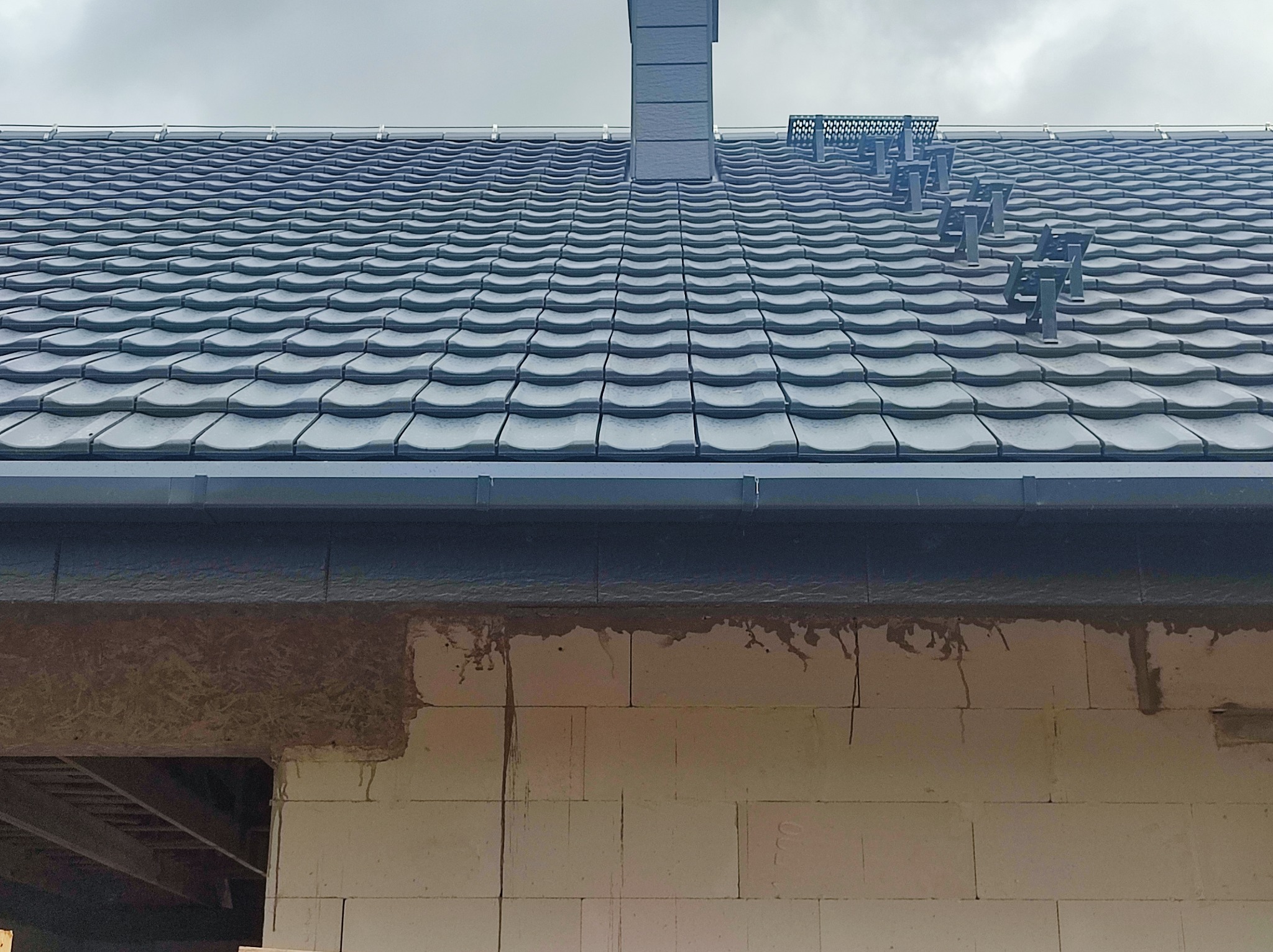 Szara dachówka ceramiczna na nowym domu w trakcie budowy, widoczne komin i stopnie dachowe, ściany z pustaków ceramicznych.