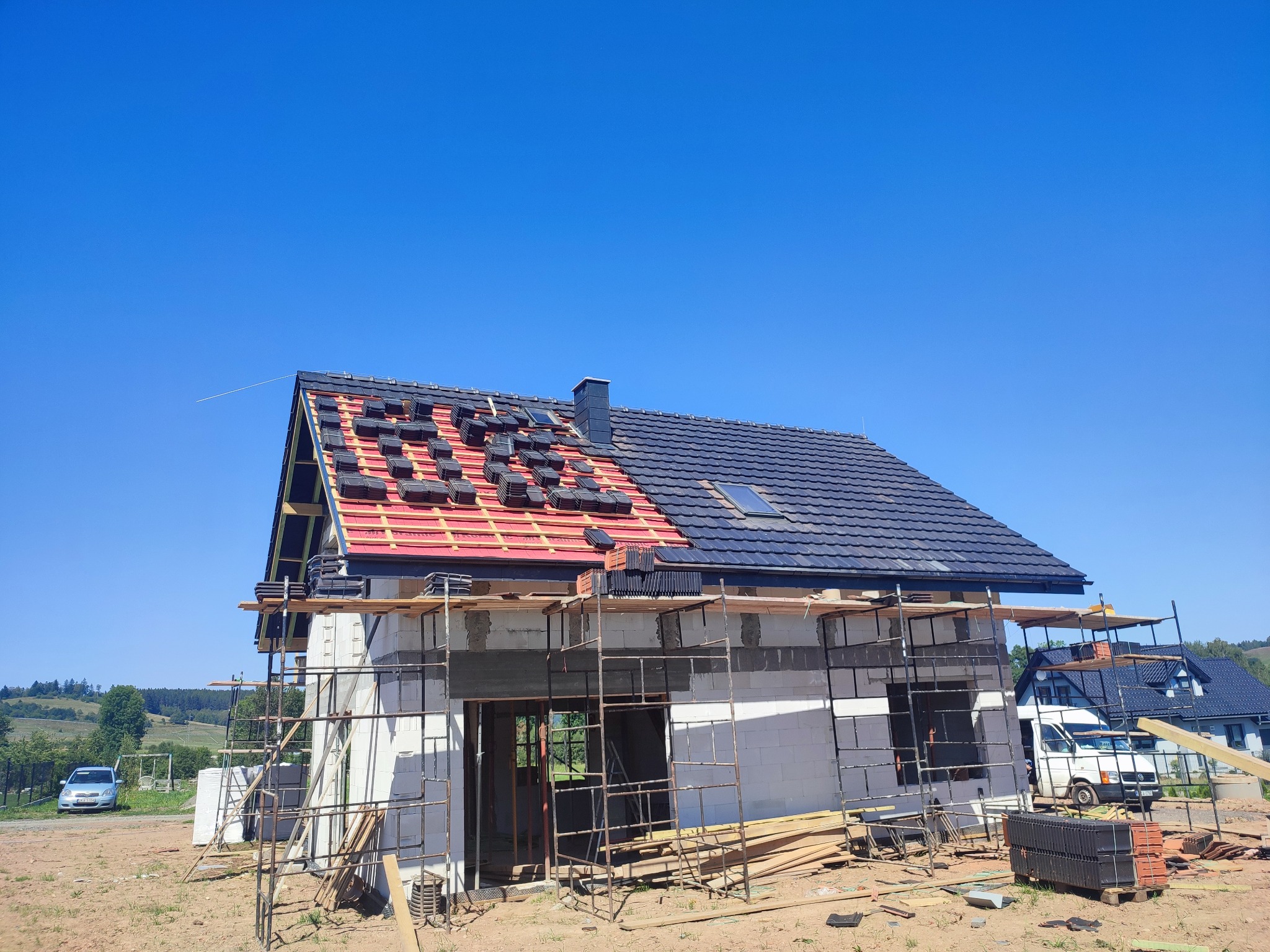 Wznoszenie domu z białych bloczków, częściowo pokrytego dachówką, z widocznym rusztowaniem i stertą dachówek na ziemi.