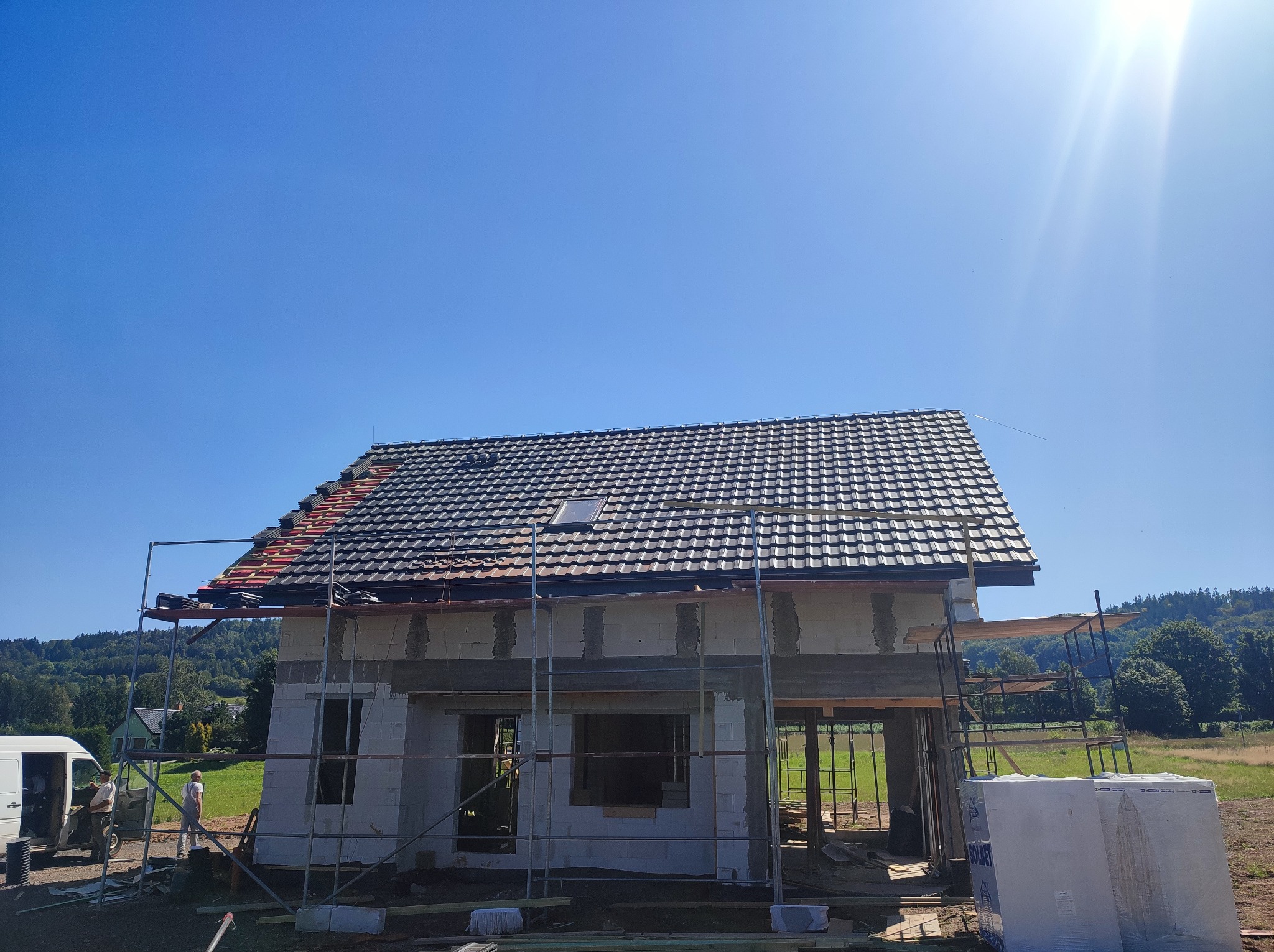 Budowa domu jednorodzinnego w trakcie realizacji: wzniesione ściany z pustaków, częściowo pokryty dach dachówką, widoczne rusztowania i materiały budowlane na tle zieleni.