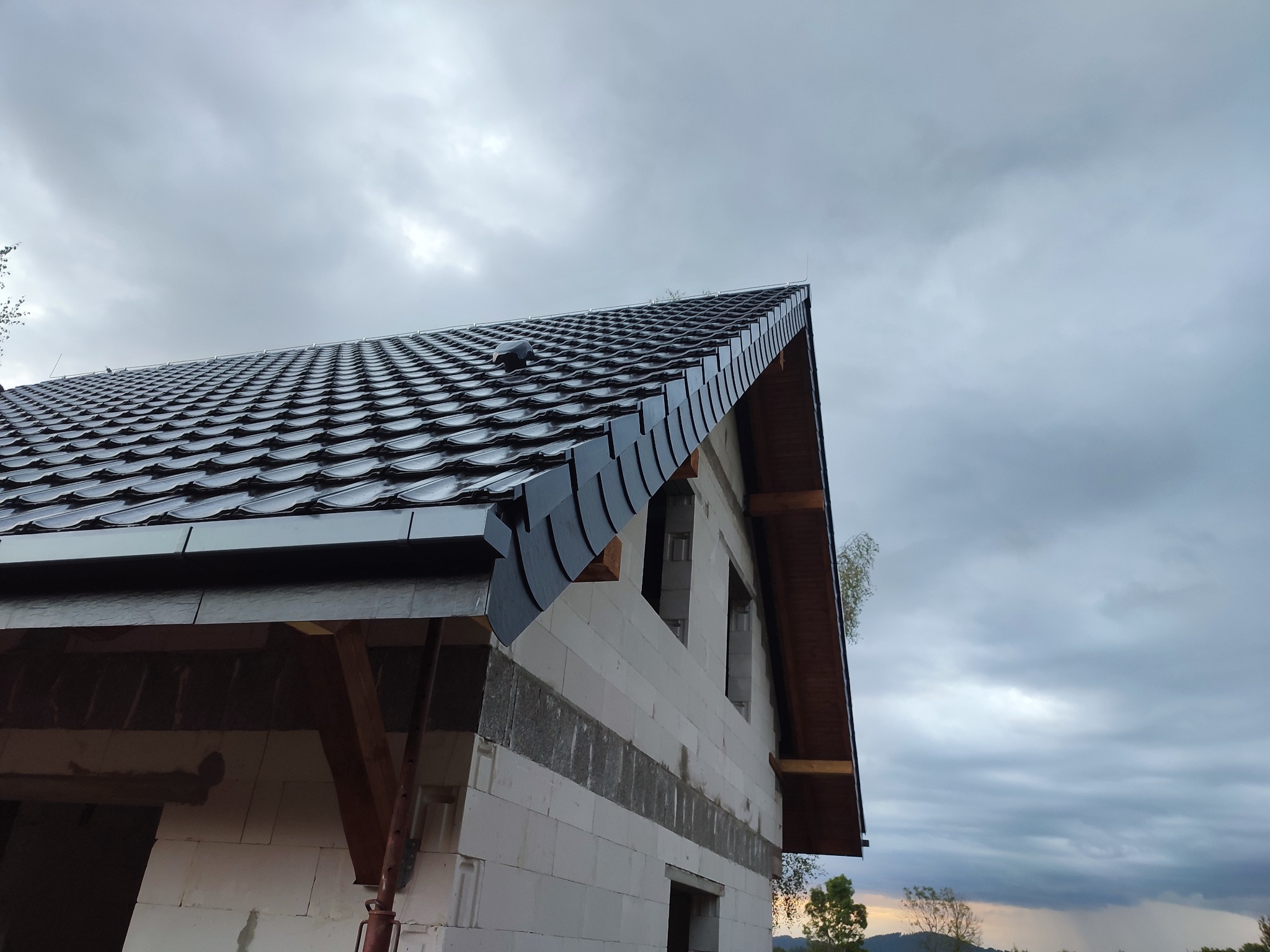 Niedokończony dom z białego pustaka z zamontowanym czarnym dachem z dachówki ceramicznej, widoczne drewniane elementy konstrukcyjne.
