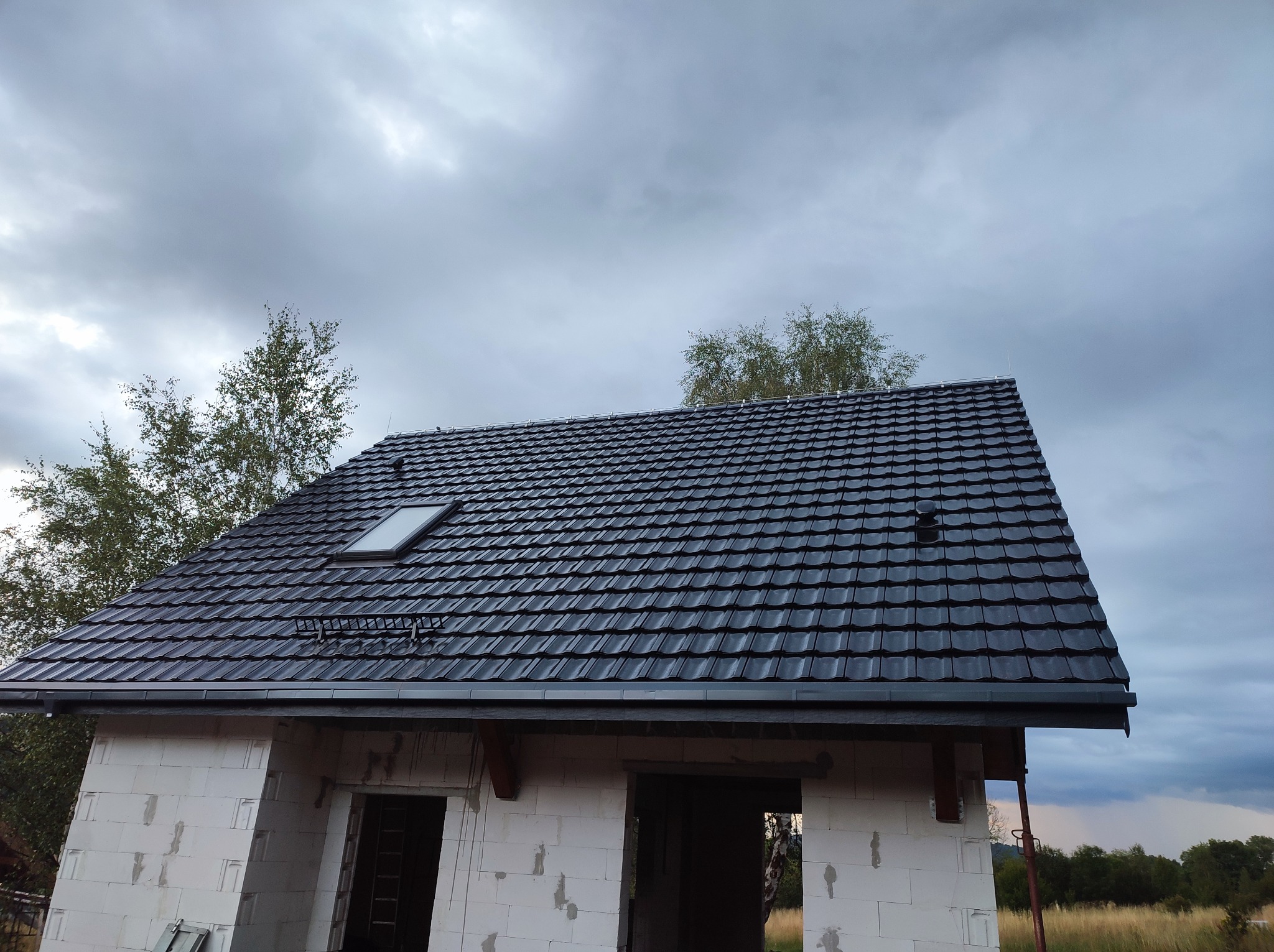 Niedokończony dom z białego pustaka z zamontowanym czarnym dachem z blachodachówki z oknem dachowym na tle pochmurnego nieba.