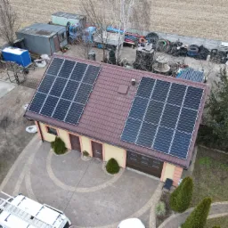 Widok z góry na dom z zainstalowanymi panelami słonecznymi na dachu, na podwórku zaparkowany biały samochód dostawczy z drabiną na dachu.