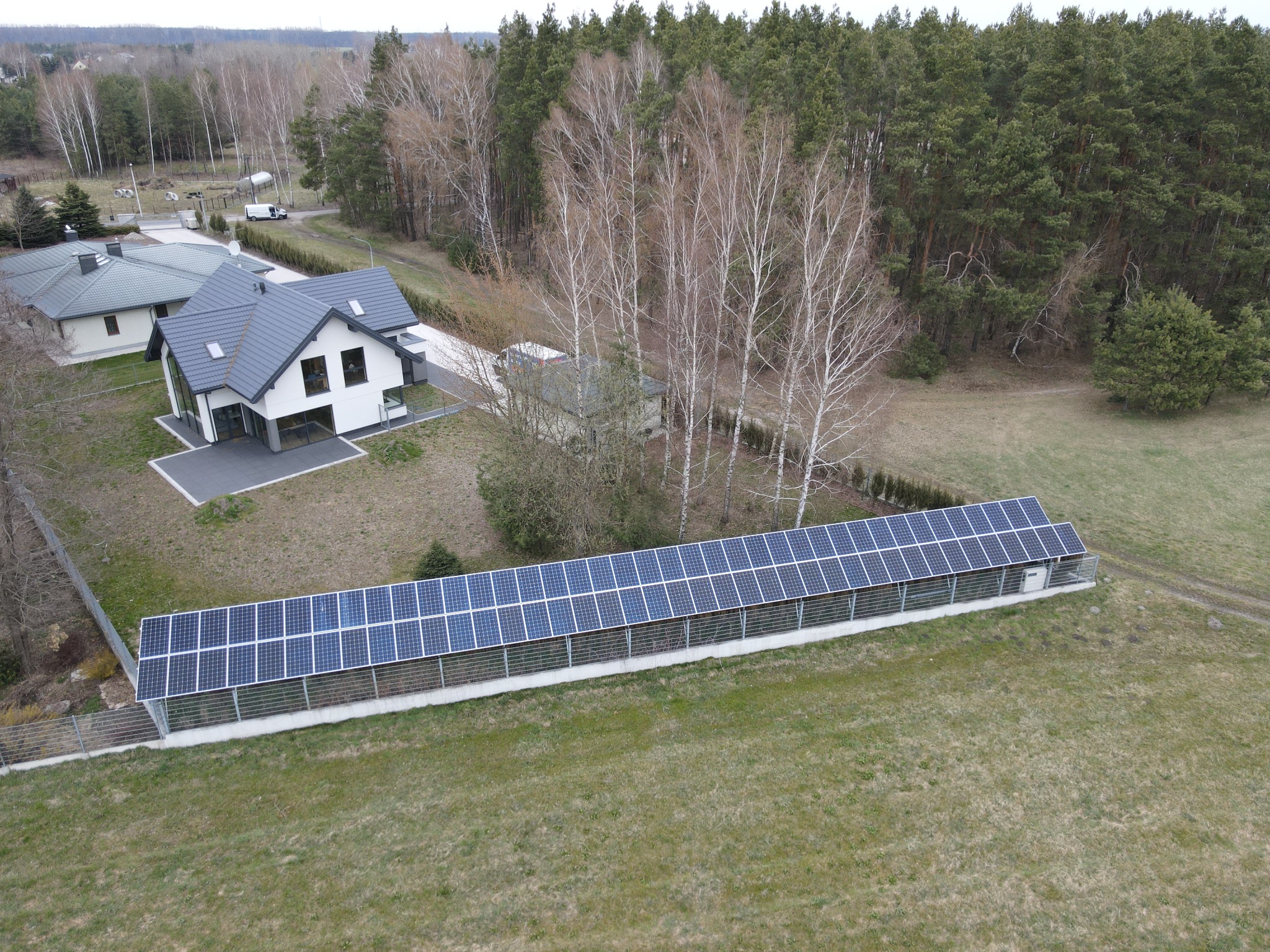 Widok z lotu ptaka na długi rząd paneli słonecznych, umieszczonych na ziemi obok nowoczesnego domu z szarym dachem i białymi ścianami, z lasem w tle.