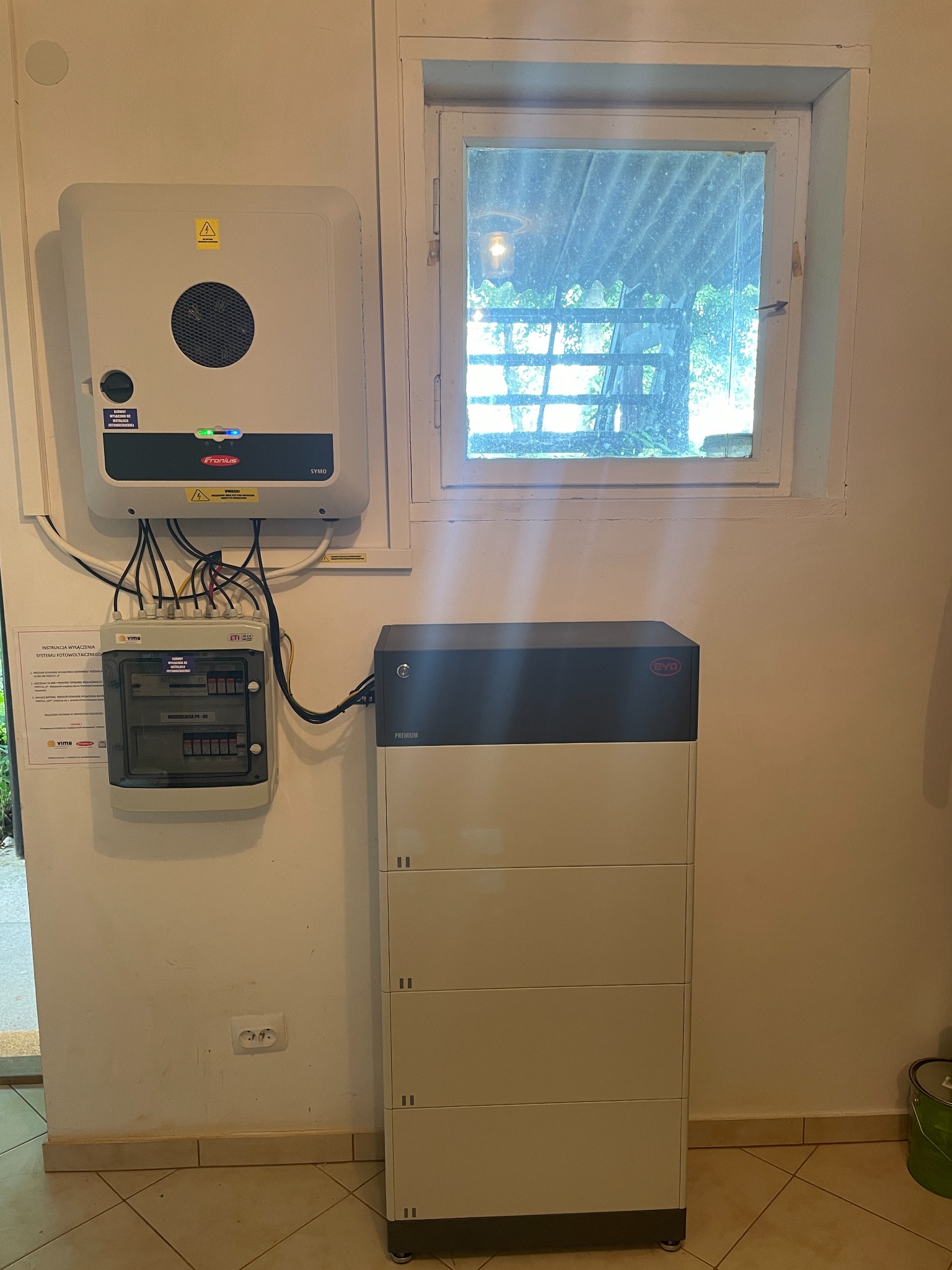 Wewnętrzna instalacja fotowoltaiczna: falownik Fronius SYMO, rozdzielnica PV-DC marki ETI oraz moduł magazynu energii BYD Premium LVL, zamontowane na ścianie w pomieszczeniu z oknem.