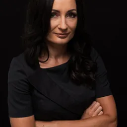 Portret kobiety prawnika w czarnej sukience, z założonymi rękami na ciemnym tle, prezentujący profesjonalizm i pewność siebie.