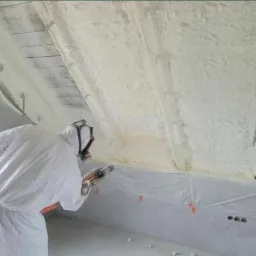 Pracownik w kombinezonie ochronnym aplikuje piankę PUR na poddasze, widoczne krokwie i zabezpieczona folią ściana, oświetlenie robocze.