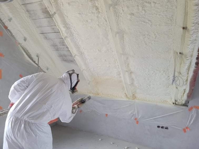 Pracownik w kombinezonie ochronnym aplikuje piankę izolacyjną na poddaszu, widoczne drewniane elementy konstrukcyjne i zabezpieczająca folia.