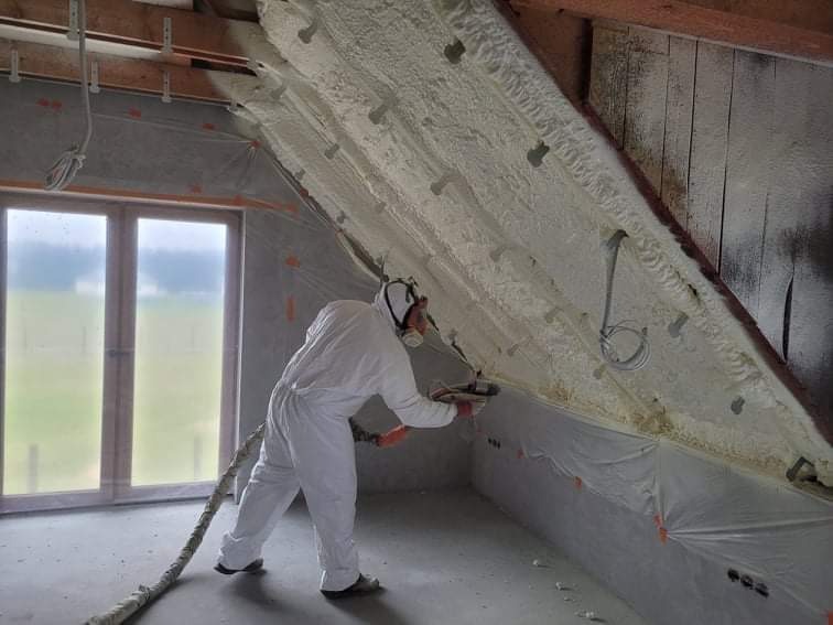 Pracownik w kombinezonie ochronnym aplikuje piankę izolacyjną na skośny sufit poddasza, widoczne elementy konstrukcji dachu i okno.