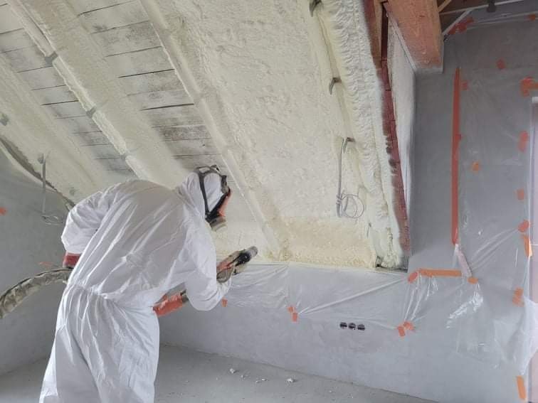 Pracownik w kombinezonie ochronnym aplikuje piankę izolacyjną na poddasze, widoczne drewniane elementy konstrukcji dachu i folia ochronna na ścianie.