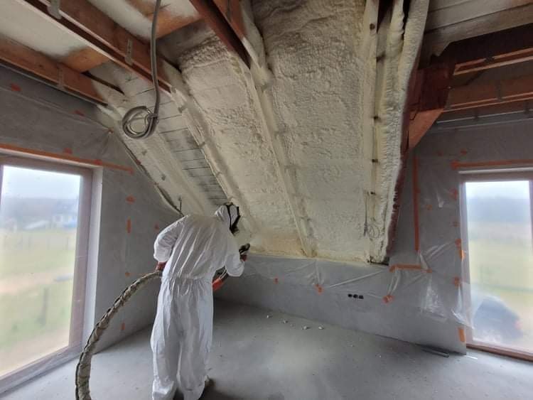 Pracownik w kombinezonie ochronnym natryskuje piankę izolacyjną na poddasze w trakcie ocieplania; widoczne drewniane elementy konstrukcyjne dachu i okna zabezpieczone folią.