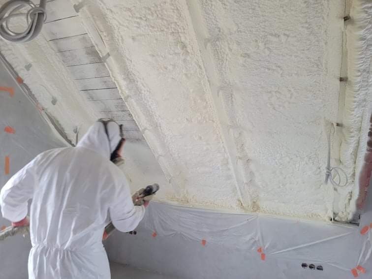 Pracownik w kombinezonie ochronnym aplikuje piankę izolacyjną na poddaszu, widoczne drewniane elementy konstrukcji dachu i zabezpieczająca folia na ścianie.