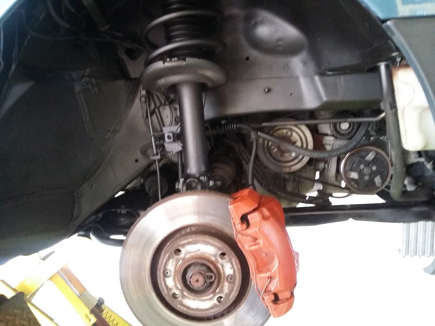 Widok podwozia samochodu z widoczną tarczą hamulcową z pomalowanym na czerwono zaciskiem, amortyzatorem i elementami silnika, podczas podnoszenia na podnośniku.