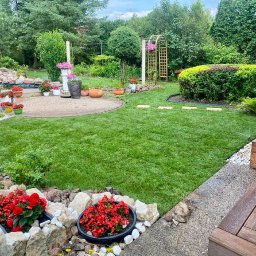 WK SERWIS - Zadbany trawnik w ogrodzie z rabatami kwiatowymi, oczkiem wodnym i elementami architektury ogrodowej. Widoczny system nawadniania trawnika w trakcie pracy.