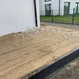 WK SERWIS - Nawadnianie piaskowego podłoża pod trawnik z rolki, w tle nowoczesny dom z ogrodzeniem. Woda rozpryskuje się z węża, tworząc efektowny wachlarz.