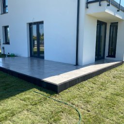 WK SERWIS - Nowoczesny taras z szarych płyt, obramowany czarną kostką, przylegający do białego domu. Soczysta zieleń trawnika i fragment węża ogrodowego w kadrze.