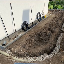 WK SERWIS - Instalacja drenażu liniowego z rurami i żwirem wzdłuż budynku w Warszawie. Widoczne narzędzia ogrodnicze i zwinięte przewody.