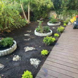 WK SERWIS - Aranżacja ogrodu z okrągłymi rabatami z obrzeżami, kamienną ścieżką i drewnianym tarasem. Widoczna gleba i świeżo posadzone rośliny.