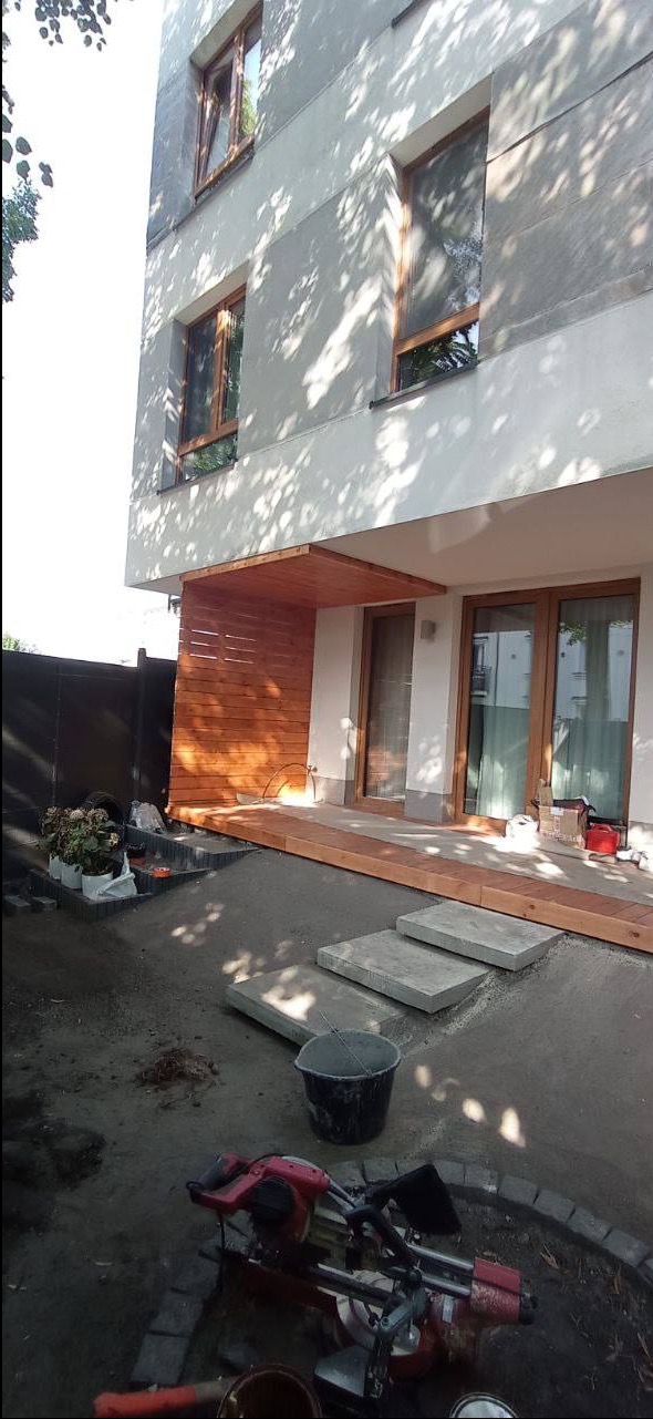 Nowoczesny taras w trakcie budowy: drewniana konstrukcja z zadaszeniem przy budynku, betonowe płyty tworzą ścieżkę, widoczne narzędzia i materiały budowlane.