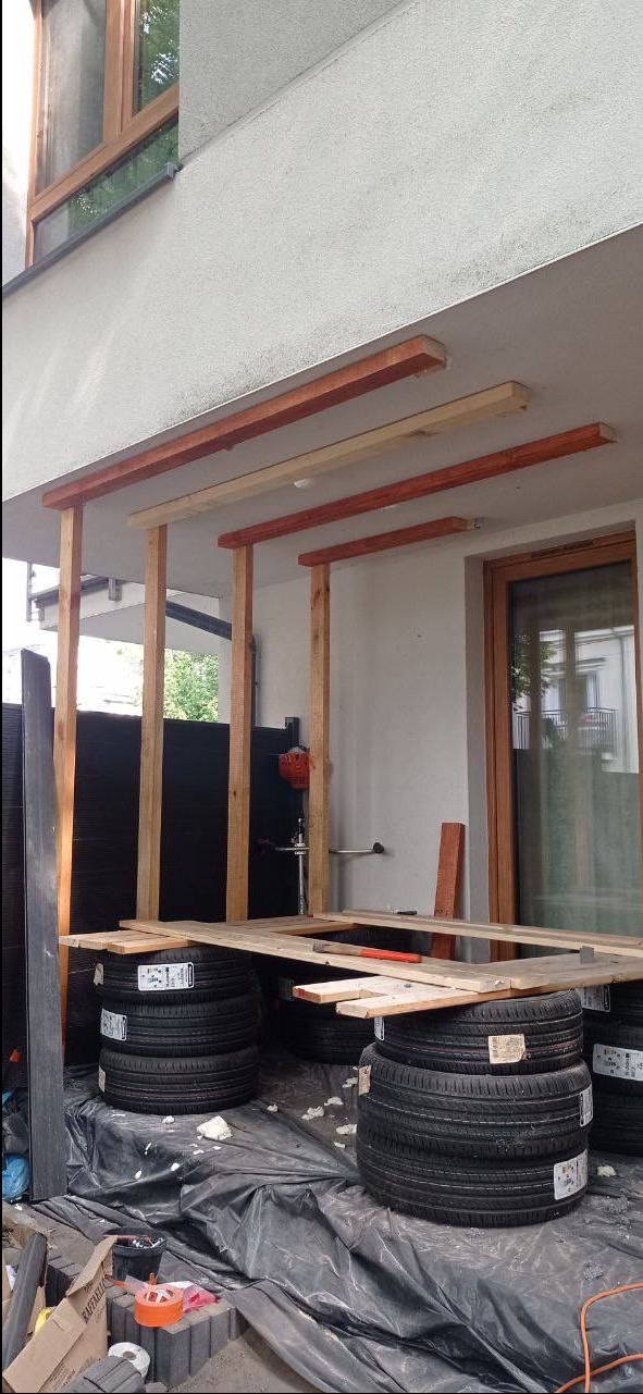 Montaż drewnianej konstrukcji z belek na tarasie, wspartej na oponach. Widoczne narzędzia i materiały budowlane. Prace w toku, surowy stan realizacji.