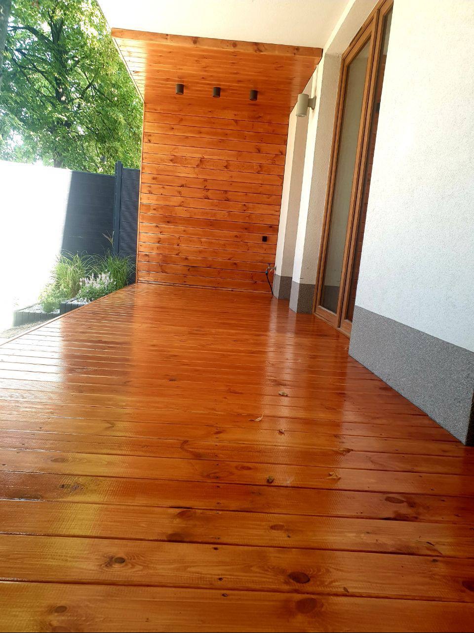 Błyszcząca, drewniana podłoga tarasu, wykonana z desek w ciepłym, miodowym odcieniu. Ściana wyłożona drewnianą boazerią. Widoczne drzwi balkonowe i fragment ogrodu.