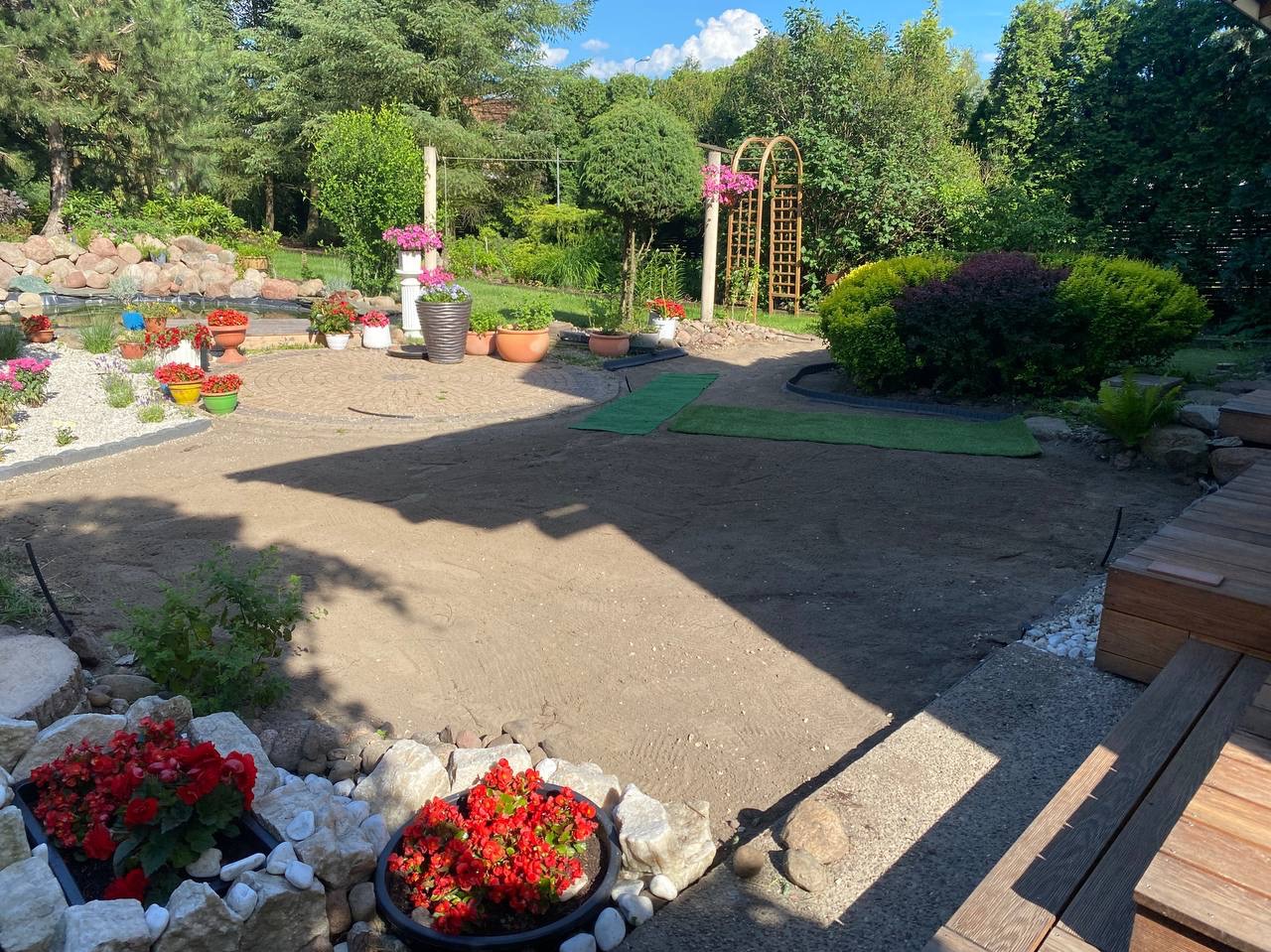 Fragment ogrodu w trakcie zakładania: przygotowany teren pod trawnik, widoczne fragmenty trawy z rolki, rabaty kwiatowe i dekoracyjny zbiornik wodny w tle.