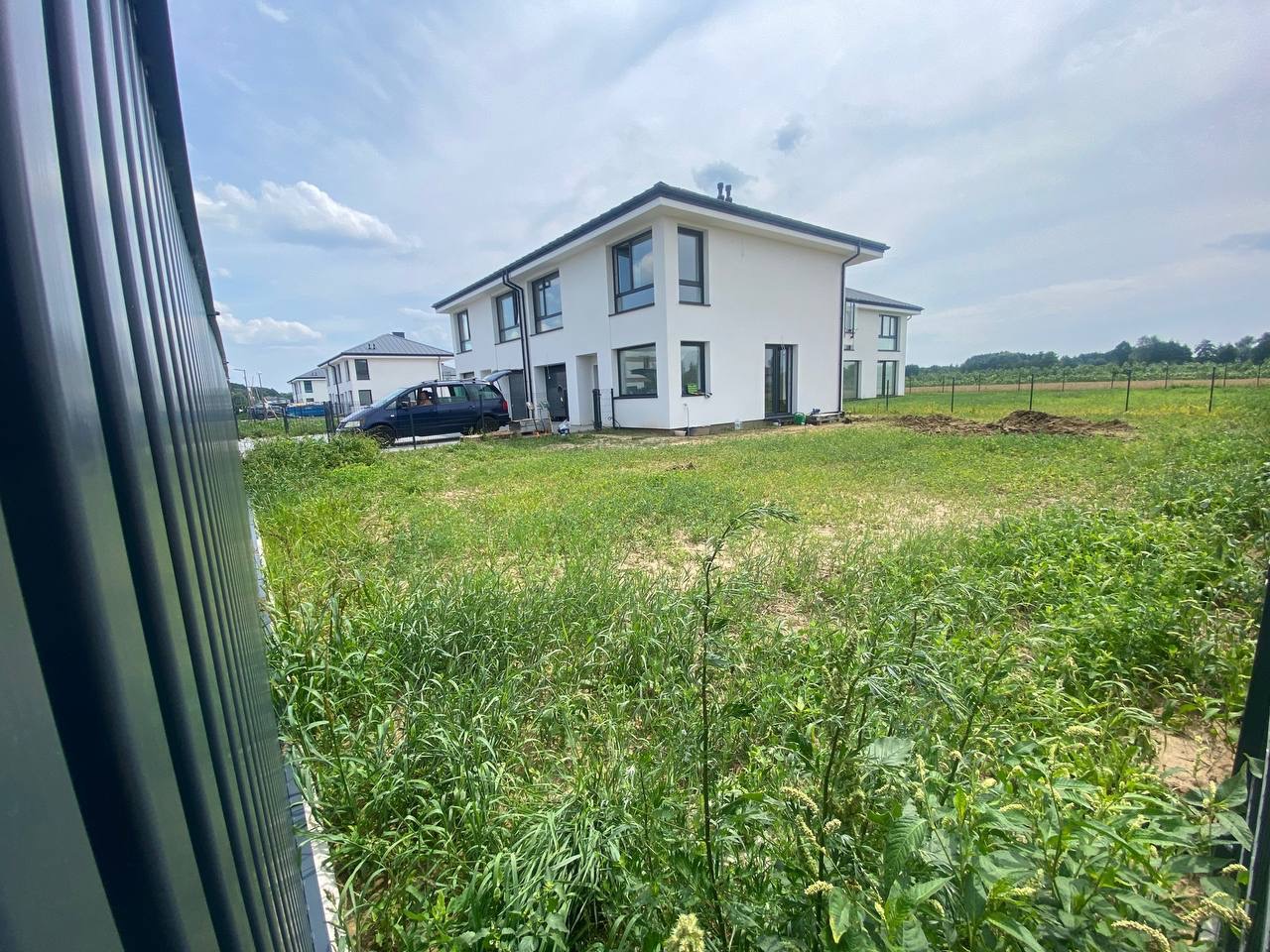 Nowo wybudowany dom z widokiem na zaniedbany trawnik i świeżo przekopaną ziemię, widziany zza ciemnego ogrodzenia panelowego.