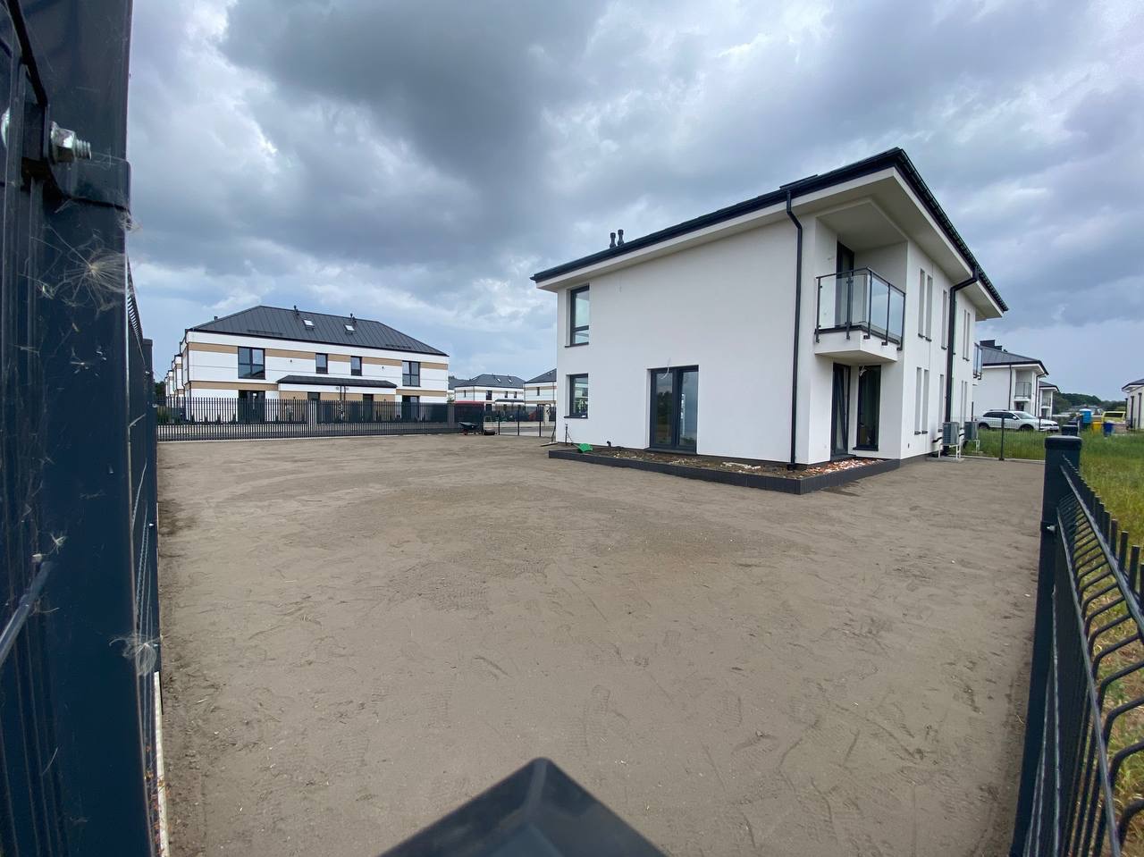 Przygotowany teren pod trawnik przy nowoczesnym domu z balkonem, widok zza ogrodzenia. Szare niebo.