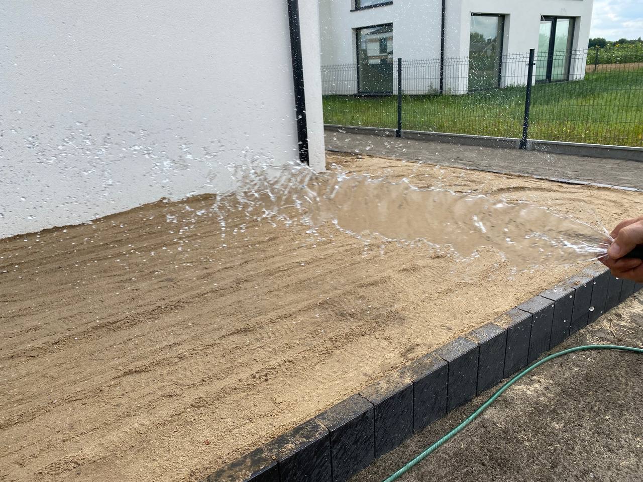 Nawadnianie piaskowego podłoża pod trawnik z rolki, w tle nowoczesny dom z ogrodzeniem. Woda rozpryskuje się z węża, tworząc efektowny wachlarz.