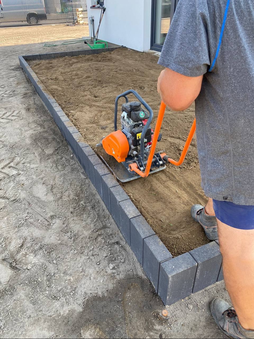 Zagęszczarka wibrująca ubija piasek w obramowaniu z kostki brukowej. Praca w toku, przygotowanie podłoża pod trawnik lub kostkę. Widoczny fragment osoby obsługującej maszynę.