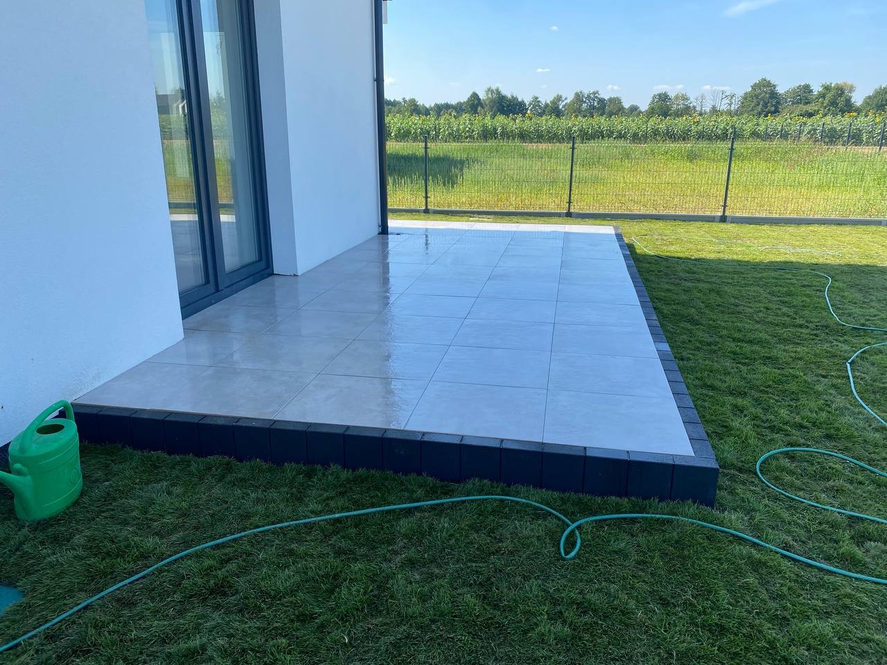 Nowoczesny taras z jasnych płytek, obrzeża z ciemnej kostki, świeżo skoszona trawa i zielony wąż ogrodowy. W tle widok na pole za ogrodzeniem.