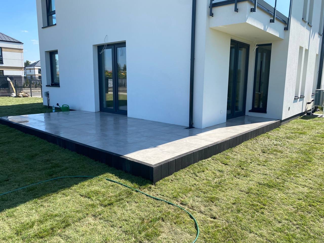 Nowoczesny taras z szarych płyt, obramowany czarną kostką, przylegający do białego domu. Soczysta zieleń trawnika i fragment węża ogrodowego w kadrze.