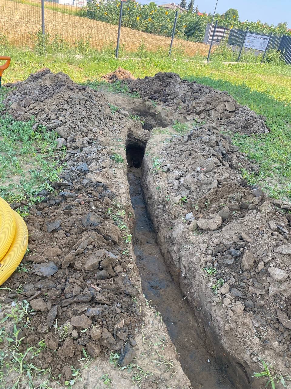 Wkopany rów w ziemi z żółtą rurą w rogu, prawdopodobnie pod system nawadniania. Widoczne pryzmy ziemi po obu stronach wykopu, w tle trawa i płot.