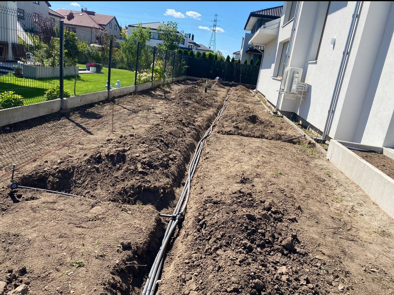 Montaż systemu nawadniania w ogrodzie: wykopane rowy z ułożonymi rurami, widok z góry, dom w tle.