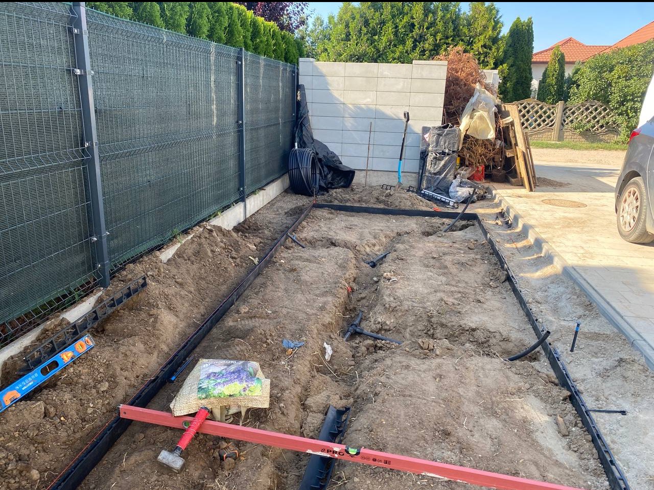 Przygotowanie terenu pod system nawadniania: wykopane rowy, rury, poziomica, narzędzia, siatka ogrodzeniowa w tle, częściowo widoczny samochód.
