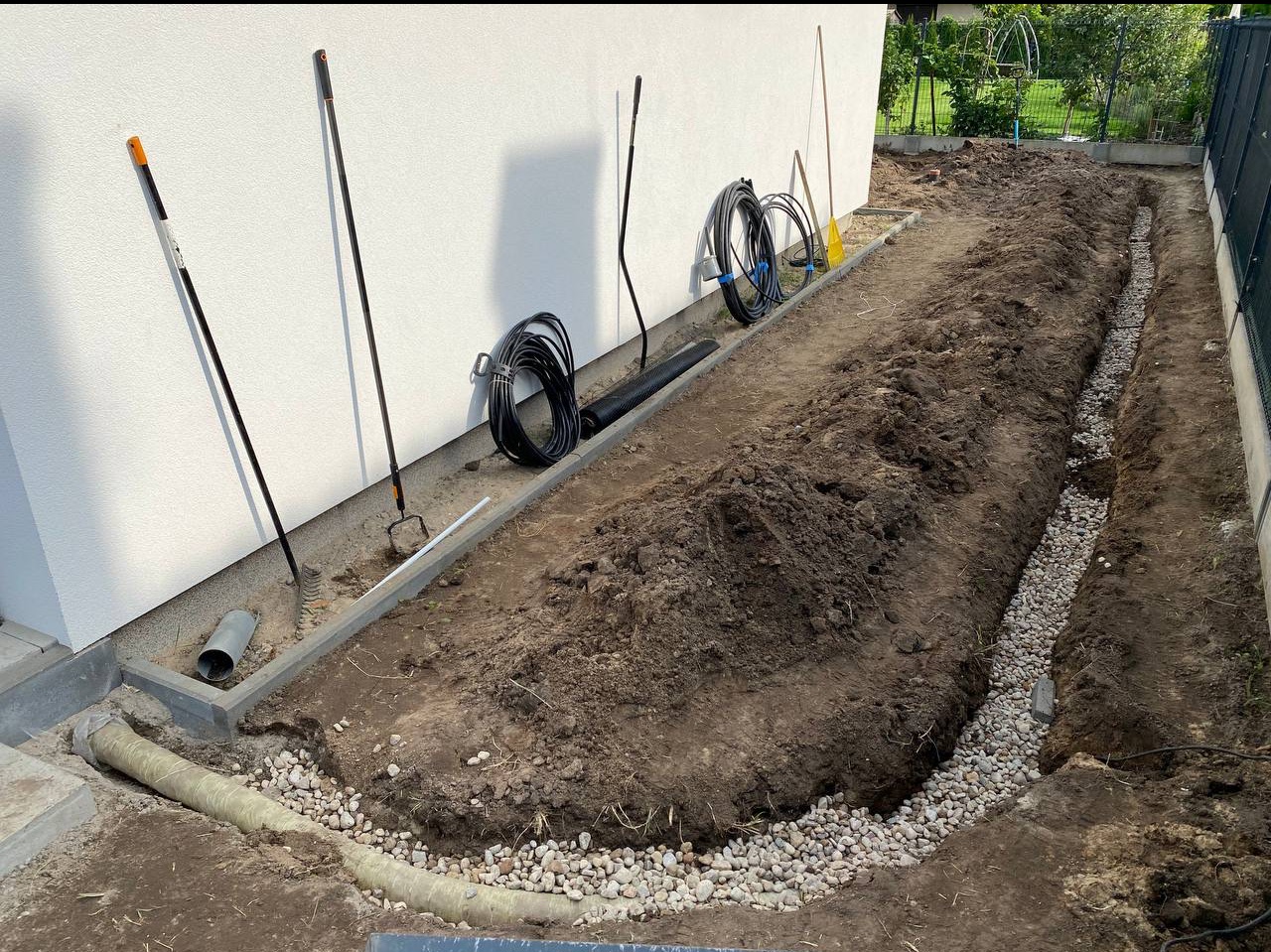 Instalacja drenażu liniowego z rurami i żwirem wzdłuż budynku w Warszawie. Widoczne narzędzia ogrodnicze i zwinięte przewody.