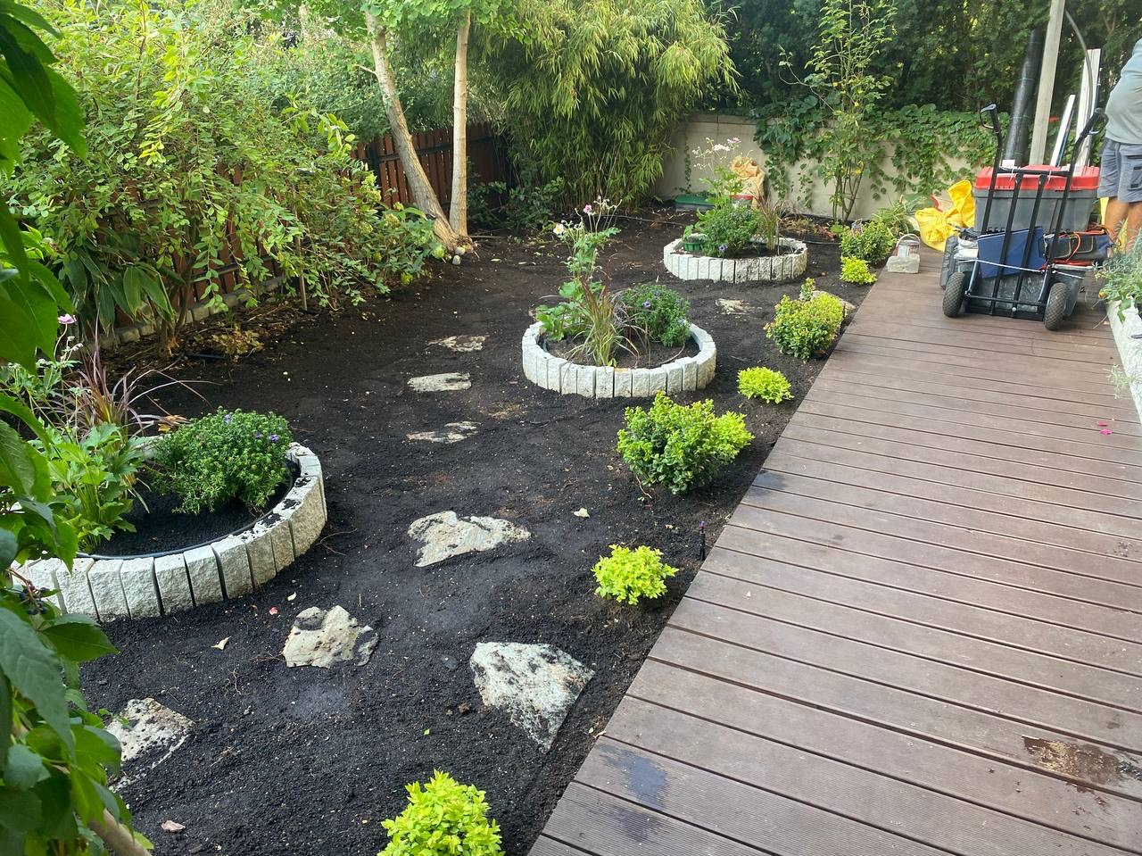 Aranżacja ogrodu z okrągłymi rabatami z obrzeżami, kamienną ścieżką i drewnianym tarasem. Widoczna gleba i świeżo posadzone rośliny.