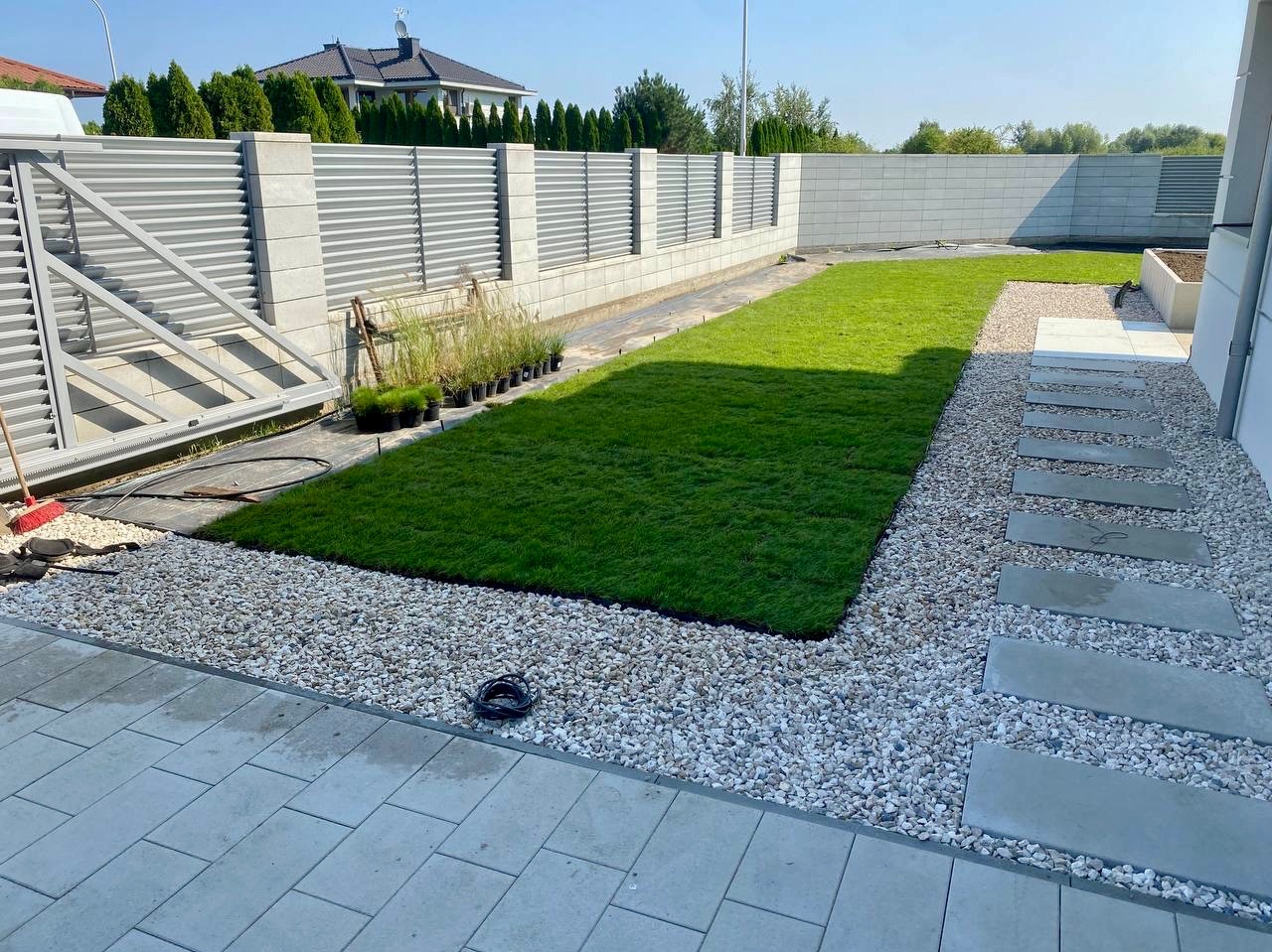 Fragment ogrodu po założeniu trawnika z rolki, widoczne obrzeża z białego kamienia, płyty chodnikowe i metalowe ogrodzenie. W tle dom jednorodzinny.