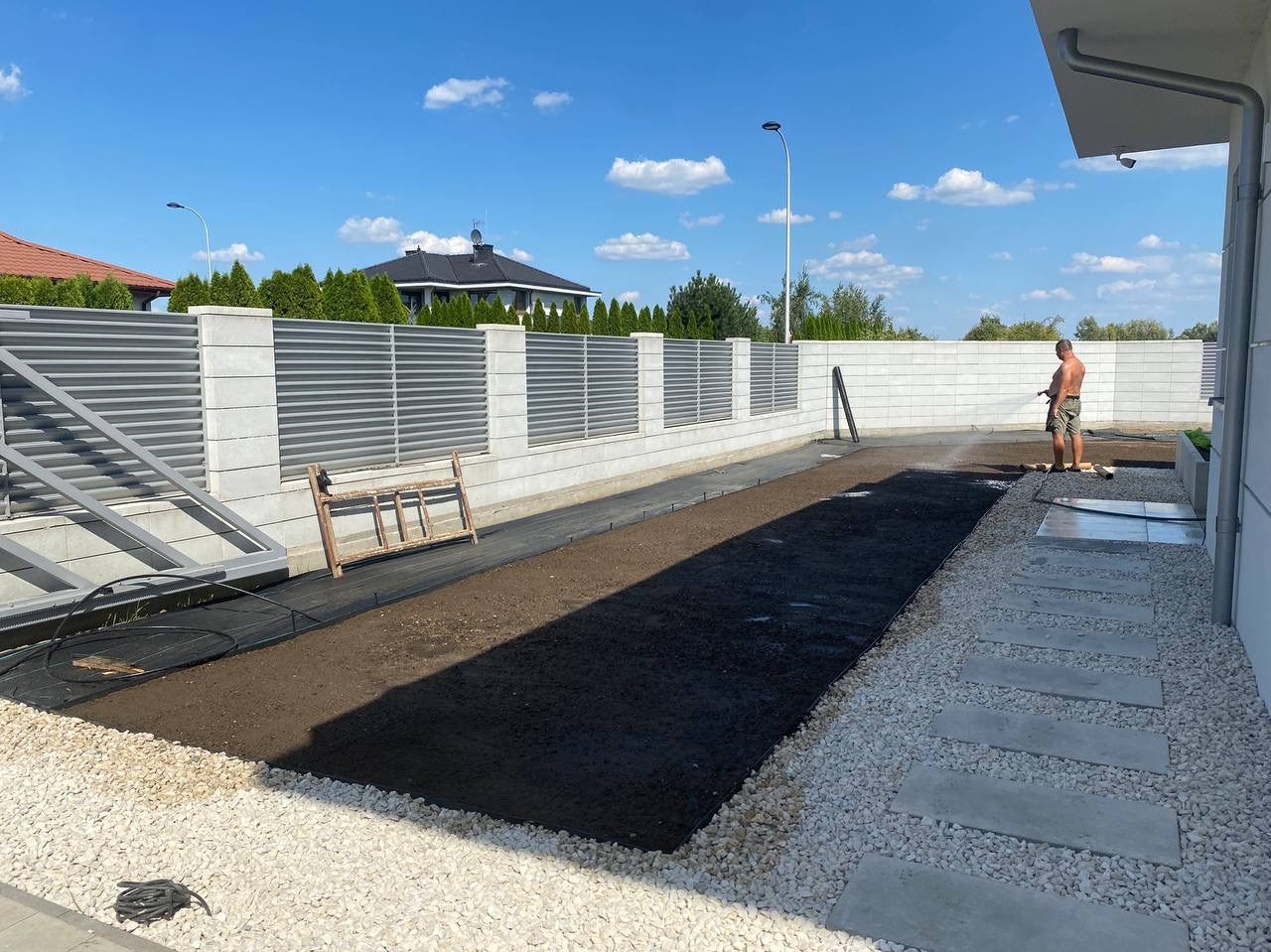Przygotowanie podłoża pod trawnik z rolki, widoczna nawodniona gleba, obrzeża z kamienia i betonowych płyt, ogrodzenie panelowe i betonowe, drabina oparta o ogrodzenie.