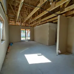 Surowe wnętrze po tynkowaniu maszynowym, widoczne drewniane krokwie dachu z instalacją elektryczną, jasne ściany z nałożonym tynkiem, betonowa podłoga z plamą światła słonecznego, okno i drzwi...