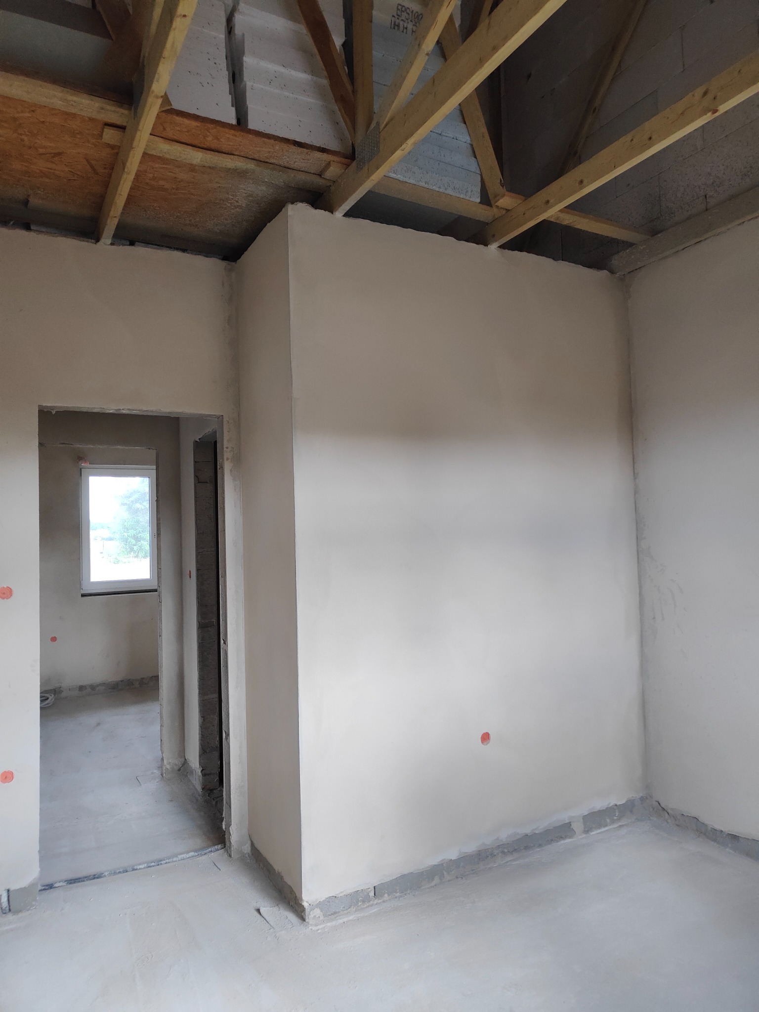 Wnętrze w trakcie tynkowania maszynowego: gładkie, jasne ściany z widocznym drewnianym szkieletem dachu, surowy stan wykończenia, przygotowane do dalszych prac.