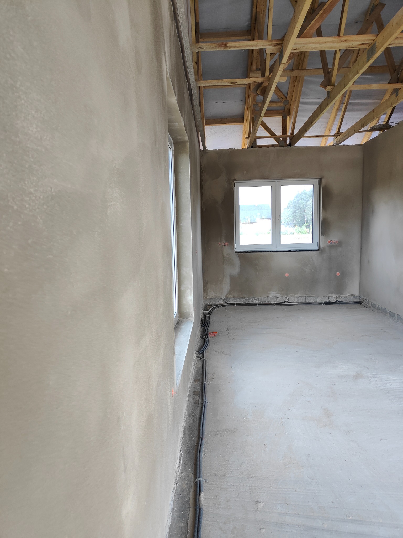 Wnętrze w trakcie prac wykończeniowych: ściany pokryte świeżym tynkiem, widoczne kable elektryczne przy podłodze, surowa konstrukcja dachu z drewnianych belek, okno z widokiem na zieleń.